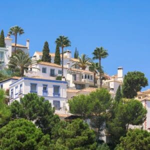 Hillside view of white Mediterranean-style houses with terracotta roofs, surrounded by green trees and tall palm trees under a clear blue sky.