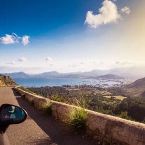 View from a car driving along a coastal road, overlooking a bay, mountains, and a small city under a bright sky with scattered clouds.