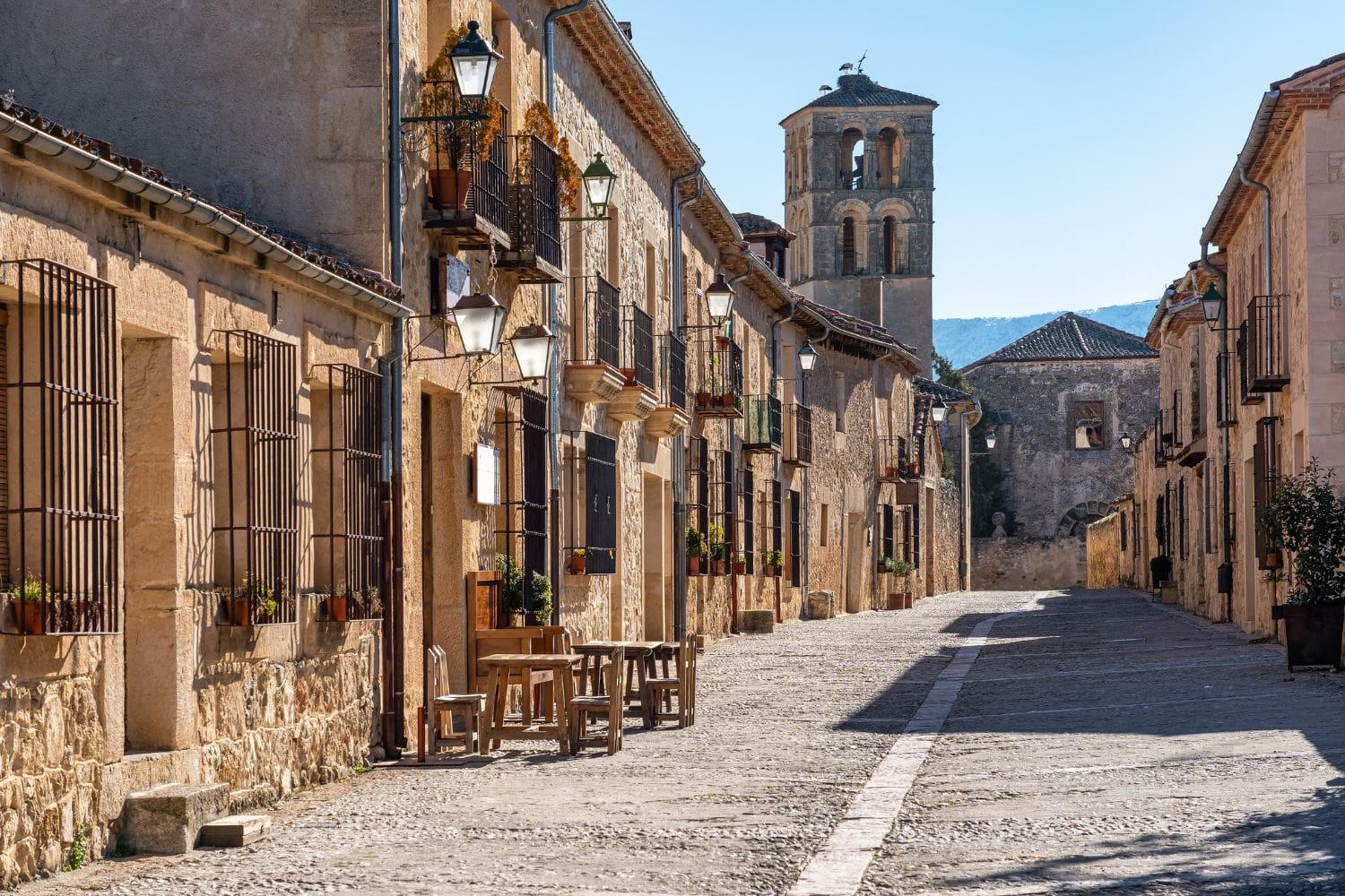 A cobblestone street lined with stone buildings and lanterns. A bell tower is visible in the background under a clear sky.