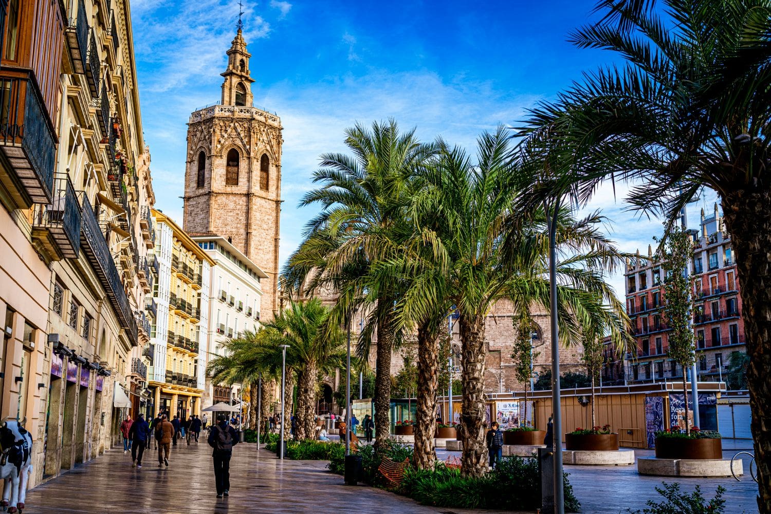 People walking along a palm-lined street with historic buildings and a bell tower under a blue sky.