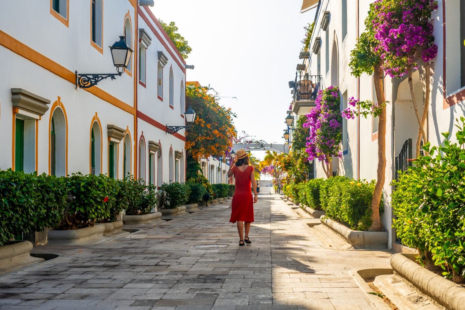Person in a red dress and hat walking down a picturesque, flower-lined street with white buildings on a sunny day.