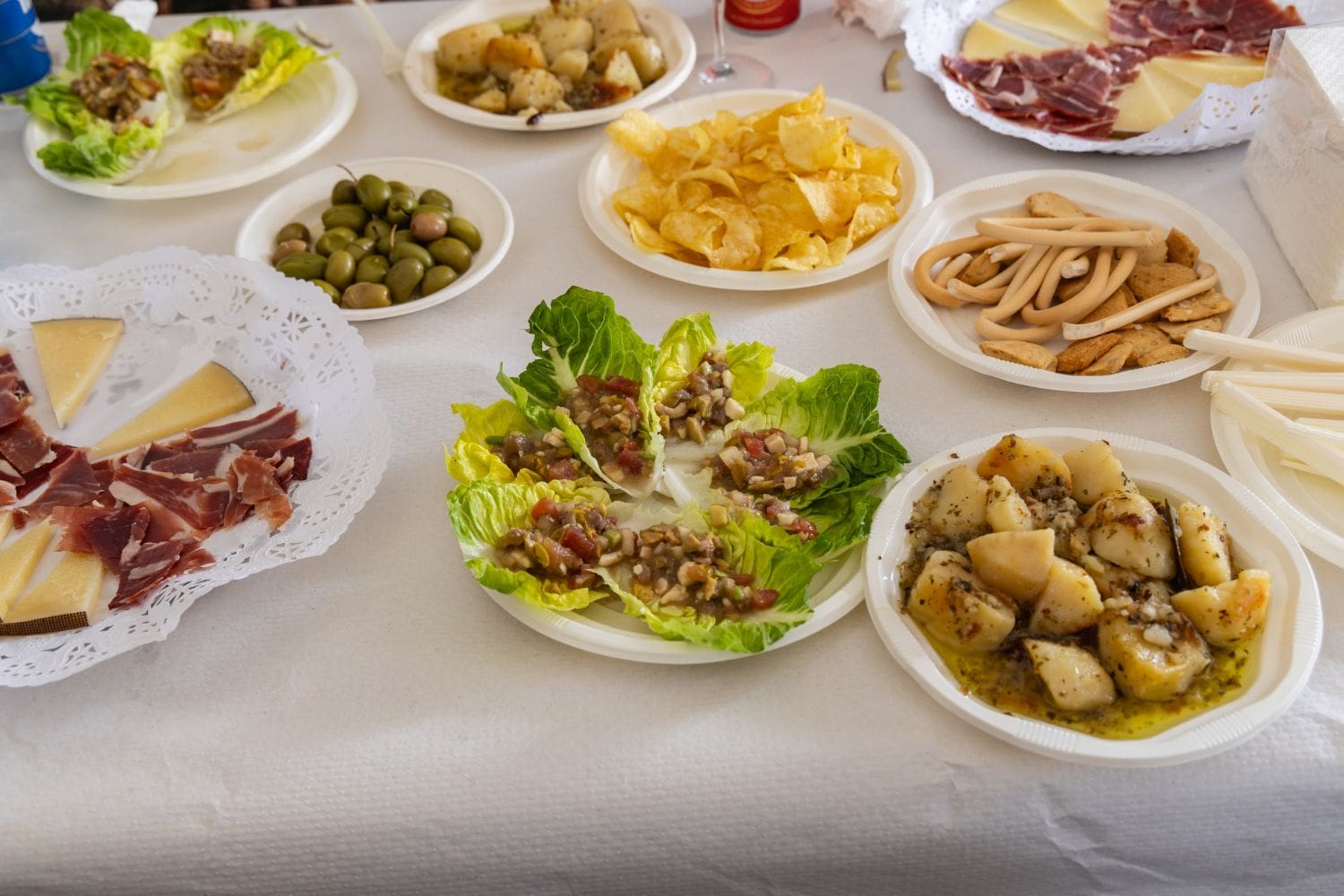 Assorted tapas on a table, including cheese, cured meats, olives, salad boats, potato dish, chips, and breadsticks on white plates.