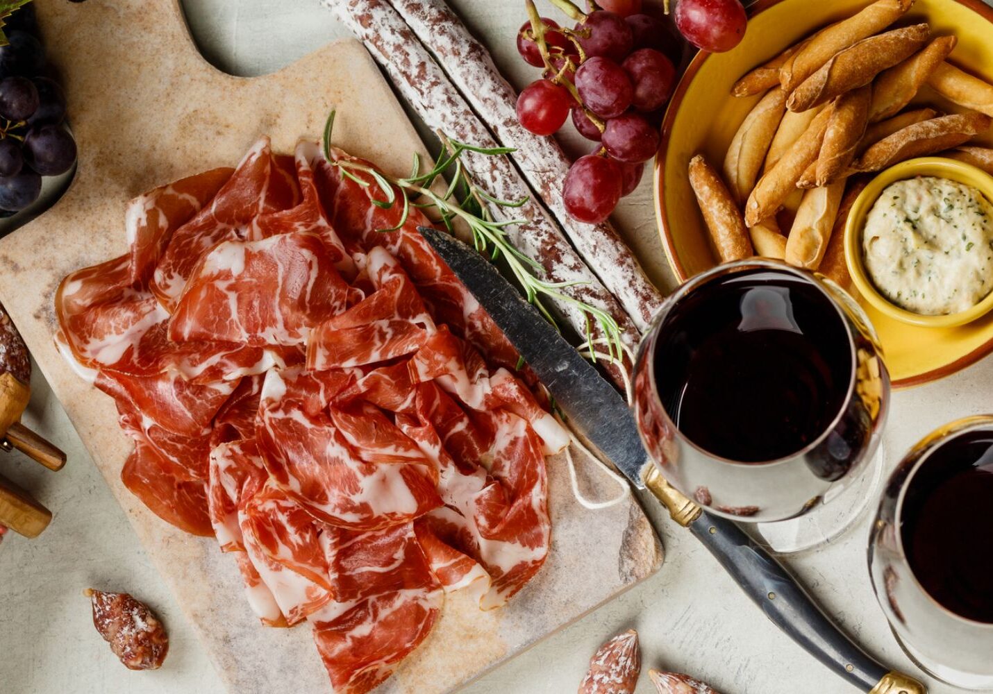 A charcuterie board with sliced cured meats, rosemary sprigs, grapes, and bread sticks. Two glasses of red wine and a plate with dip are also present.
